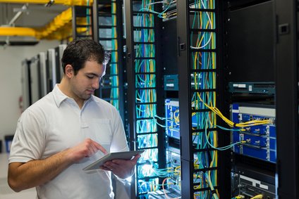 טכנאי בחדר שרתים, עומד מול ארונות תקשורת צבעוניים ומשתמש בטאבלט.