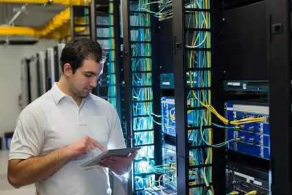 טכנאי בחדר שרתים, עומד מול ארונות תקשורת צבעוניים ומשתמש בטאבלט.
